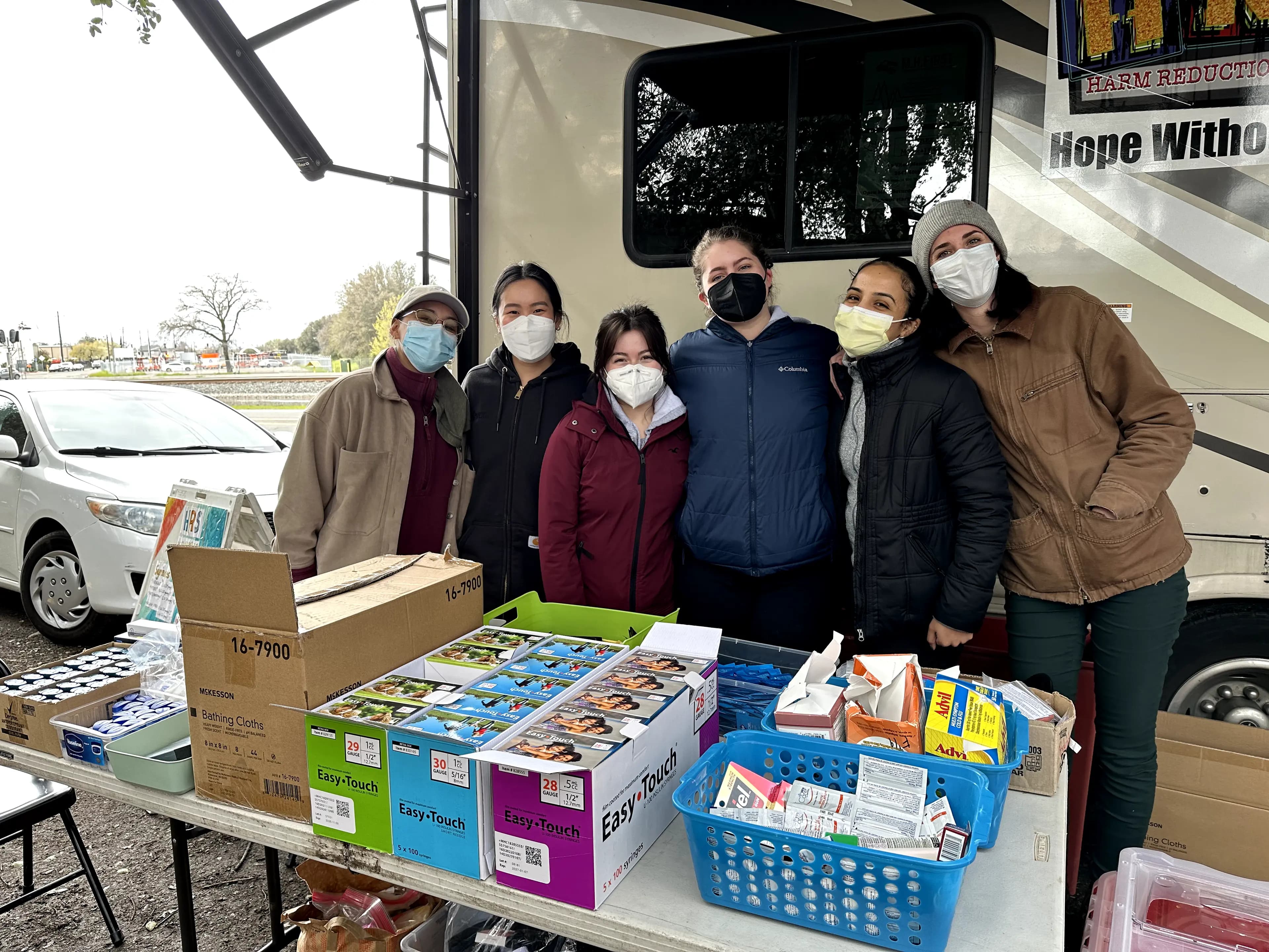 A group of people wearing masks dressed warmly in jackets and coats. They are standing outside, behind a table aligned with different boxes of vaseline, insulin syringes, and over the counter medications.