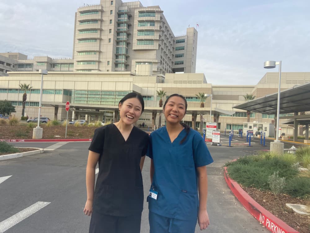 Two female doctors, wearing scrubs, are smiling and posing in the parking lot in front of the Sutter Health building during a volunteering event. 
