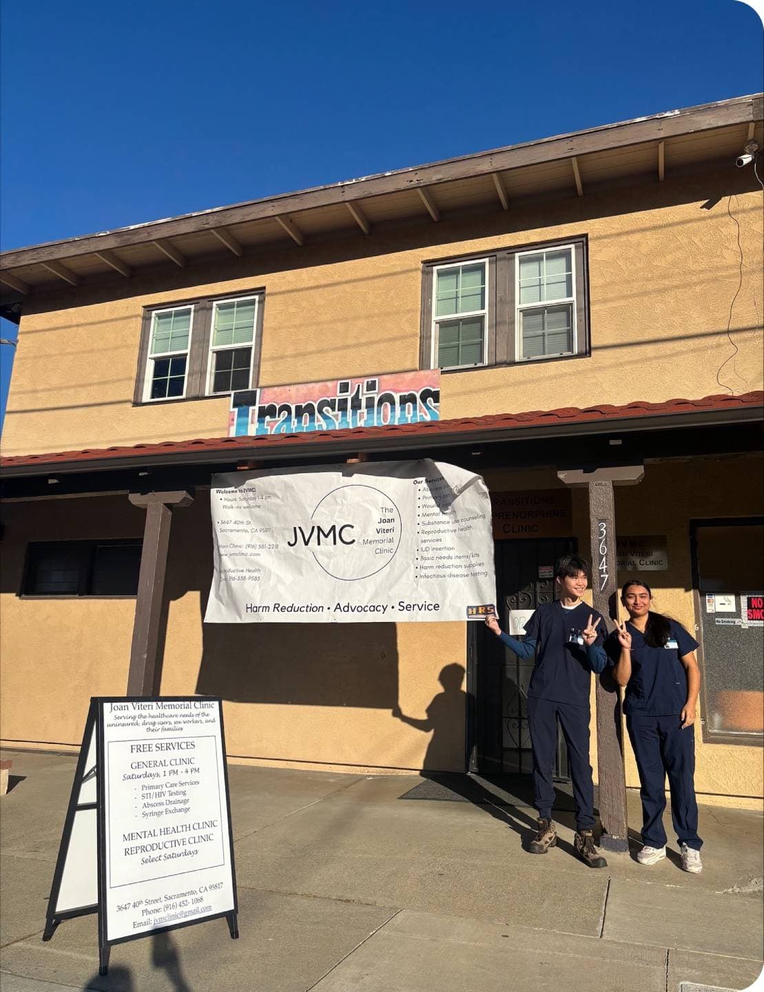 A male and female students, in scrubs, stand in front of the JVMC building smiling and posing. There is a white banner with the JVMC logo on it hung from the awning. A sign outside of their door states the services the clinic provides. 
