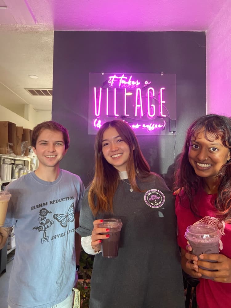 Three people are standing together in front of a neon pink sign that reads "It Takes a Village (and a cup of coffee)." They are holding iced drinks and wearing casual clothing, including shirts supporting harm reduction programs.