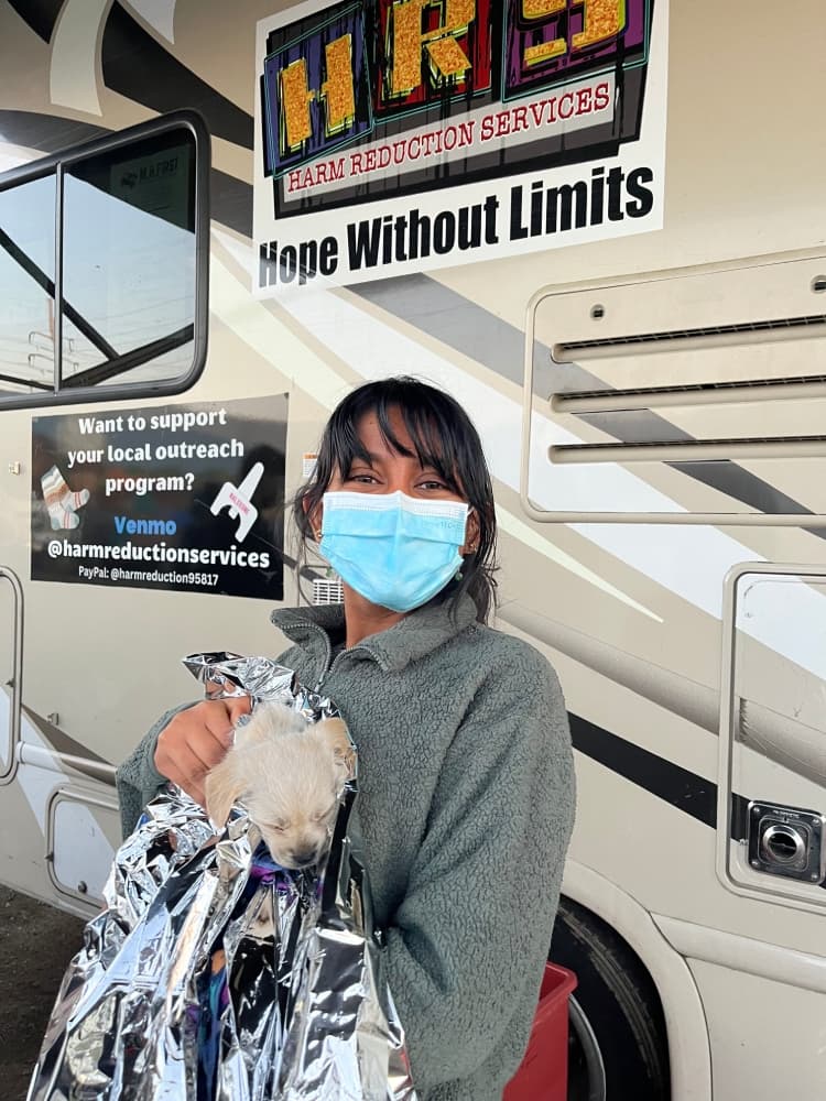 A person wearing a blue face mask and a gray fleece jacket holds a small, light-colored puppy wrapped in a reflective emergency blanket. They are standing in front of a harm reduction services vehicle with signs promoting outreach support and the slogan "Hope Without Limits."