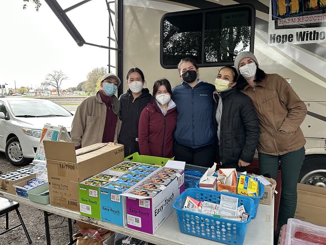 A group of women wearing masks dressed warmly in jackets and coats. They are standing outside, behind a table aligned with different boxes of vaseline, insulin syringes, and over the counter medications.