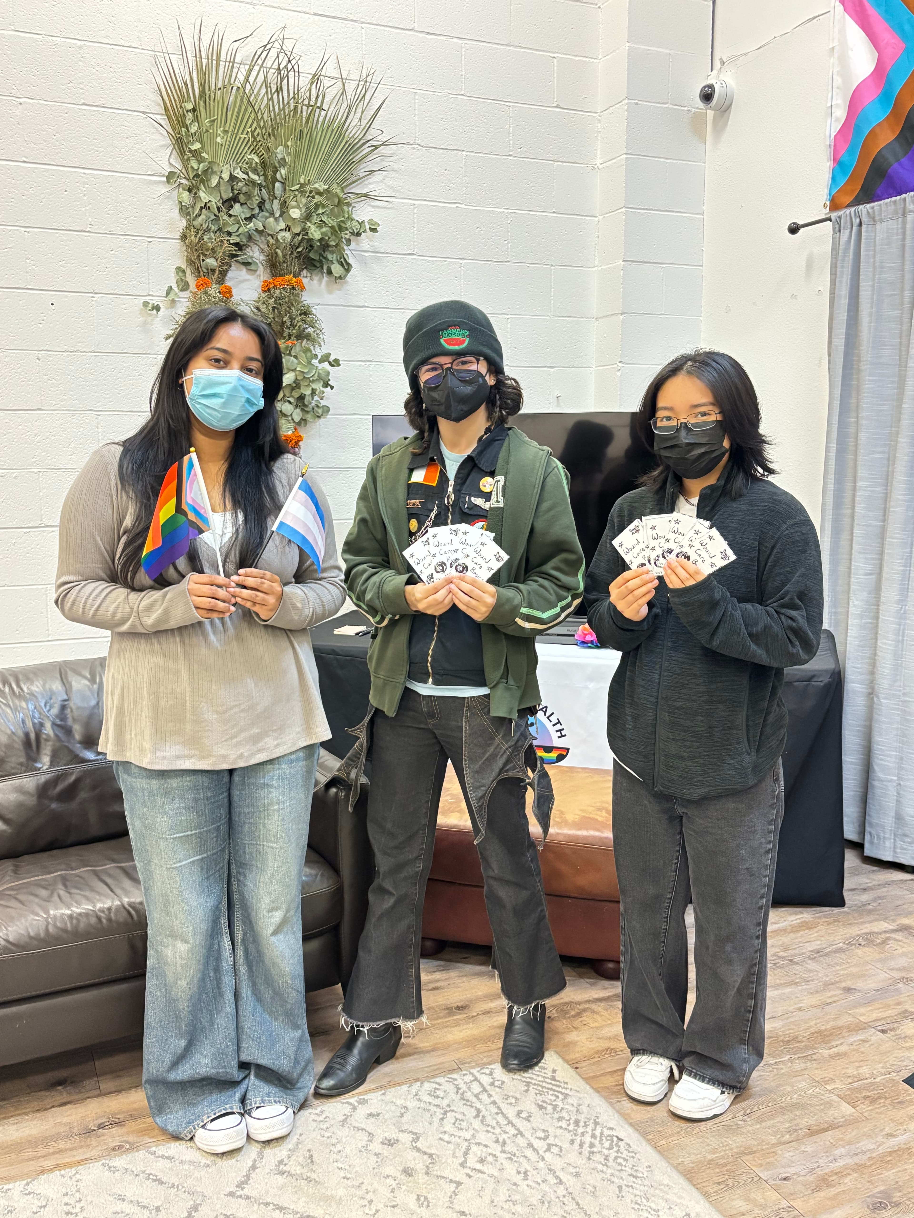 Three women are standing holding various things. The woman on the left is holding various small LGBTQ flags. The two women on the right are holding up many cards that read "Wound Care Basics."
