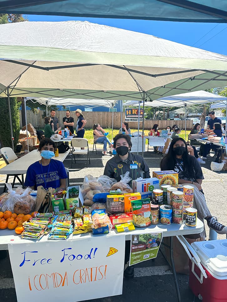 Three people sit at a tent booth behind a foldable table which has a poster taped to the front that reads: "Free Food! Comida Gratis!" The table is stocked with a variety of food items such as cereal, protein bars, fruit, canned goods, and bread.