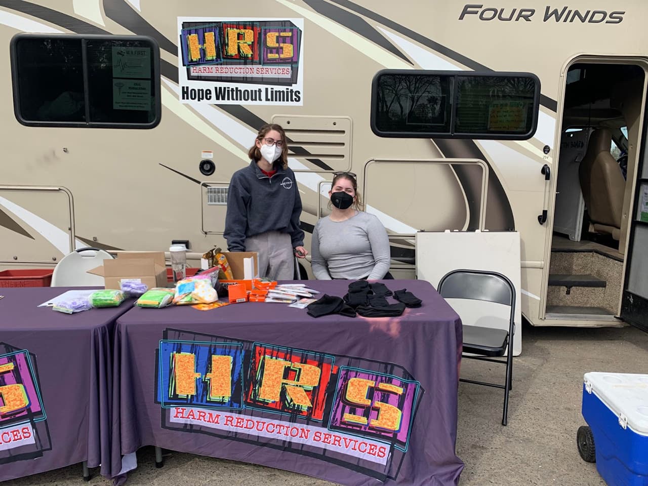 Two people are behind a table covered with purple table cloth that says "HRS Harm Reduction Services" in large letters in the front, and on top of the table lies essential items such as black socks and pads. The table is set up in front of a large tan van, which has a poster taped to it reading, "HRS Harm Reduction Services. Hope Without Limits." 