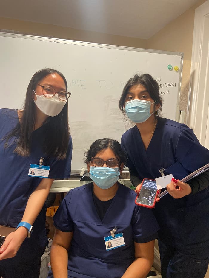Three women wearing navy blue scrubs, masks, and name tags pose in front of a whiteboard. One is holding a tablet and another is holding a laptop. 
