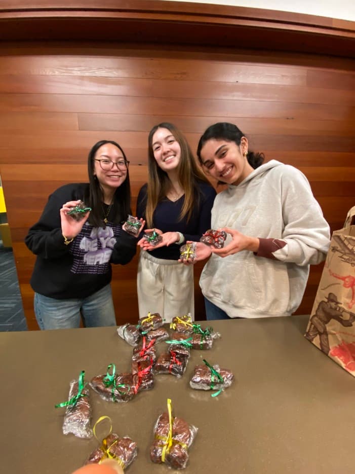 Three women standing next to one another, each holding a plastic bag with a brownie inside tied with a red, green, or yellow ribbon. The three girls are all standing behind a table with more brownies scattered on top.