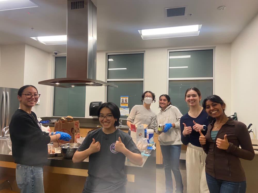 Six college girls baking goods in a kitchen while volunteering at Joan Viteri Memorial Clinic. The girl on the the far left is cracking eggs in a bowl while the other five girls surrounding the table are giving a thumbs up and a smile. 