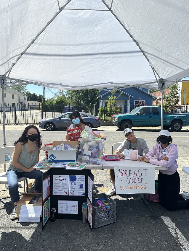 Four female students are sitting at a table educating the public about breast cancer screenings, a service their clinic provides. There is a black trifold poster board presentation in front of table. A breast cancer screening sign is taped to the front of the table. A lot of bins full of supplies are on the table. 