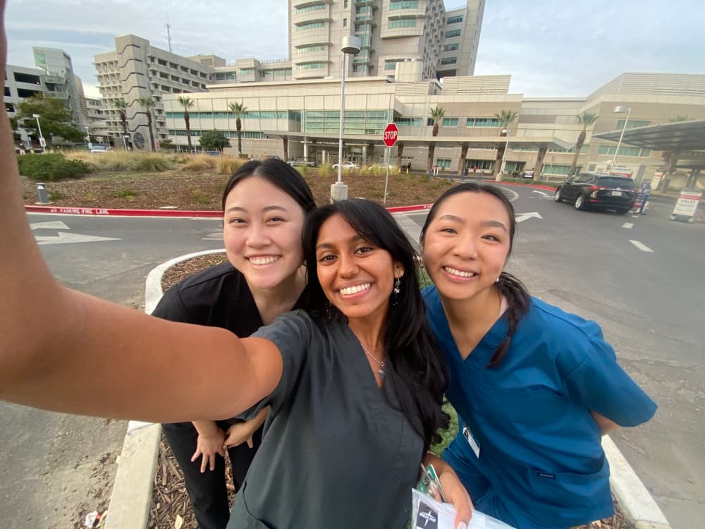 Three girls taking a selfie in front of hospital. All three girls are smiling and wearing different colored scrubs; from left to right, black scrubs, grey scrubs, and blue scrubs. 
