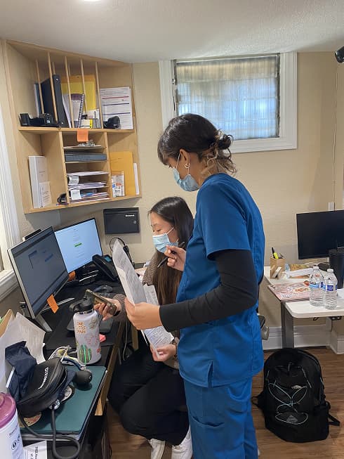 Two female doctors, wearing face masks, are looking at the computer in an office in the JVMC building. One doctor is standing up with papers and a pen in her hands and the other doctor is sitting down with papers and her phone in her hands. 